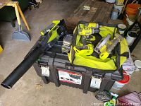 Overview photo showing all Ryobi tools including blower, drills, circular saw, flashlight, battery charger, battery, bits, blade, and tool bag arranged on and around a black plastic toolbox in a garage setting.