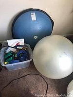 Photo of blue Bosu balance trainer, silver Fitball, crate holding various weights, DVDs, and workout book on carpet.