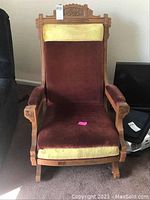 Front view of antique Eastlake platform rocker chair showing wood carvings and two-tone upholstery with burgundy seat and gold accents on headrest and top back cushion.