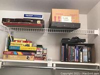 Boxes of board games and puzzles stacked on wire shelving, several labeled boxes visible including Dominoes, Scrabble, and Perquackey, along with DVDs and a DVD player with remote.