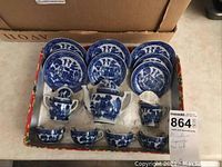 Miniature Japanese tea set in original box featuring teapot, sugar bowl, creamer, cups, saucers, and small bowls with blue and white porcelain designs. One cup has broken handle.