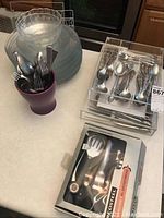 Overview of glass plates and bowls stacked, purple vase containing flatware, two plastic cases with flatware, and boxed corkscrew set on countertop.