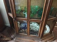 Wide shot showing two shelves of glassware and china items inside a wooden glass-front cabinet.
