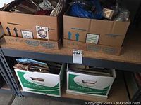 Two cardboard boxes on a metal shelf containing various craft materials including yarns, fabric, and paper flowers.