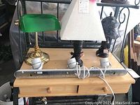 Four table lamps placed on a wooden surface showing varied styles and colors including green glass shade, white fabric shade, stained glass style shade, and horizontal lamp fixture.