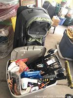 Photo showing the wheeled suitcase leaning against a shelf and a plastic bin filled with household items including cleaning brushes and various personal care products.
