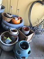Photo showing large gray planter with soil and rods, ceramic, plastic, and terracotta pots inside and around, along with a wooden planter box with wheels and a metal garden decor piece behind.