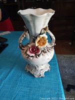 Ivory white Capodimonte floral porcelain vase with decorative raised roses in yellow and pink, sculpted scrolled handles and ornate pedestal base, photographed on blue textile surface with wood furniture background.