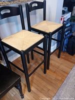 Two black wooden stools with square twine wrapped seats and cutout handle backs, positioned side by side on a hardwood floor.