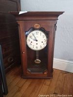 Full view of wooden wall clock with glass front and visible pendulum on floor near wall.