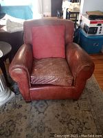 Photo showing brownish red leather armchair with a red fabric pillow placed on the seat against the backrest, highlighting the wear and scratches on the leather, and overall size and shape of the chair.