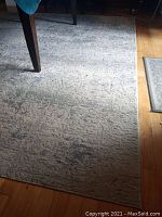 Close-up photo of the rug under a table showing the texture and color pattern in white, grey, and some blue hues. Hardwood floor is visible around the rug.