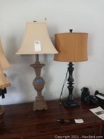 Front view of two pedestal lamps on a wooden surface against a wall, showing gold lamp with beige square shade and black lamp with brown round shade.