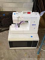Full view of white sewing machine sitting on top of white GE microwave oven against a concrete floor background.