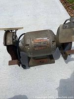 Full front view of the gray metal BA Forge model HG6 bench grinder showing both wire wheel and grinding wheel sides, with protective guards and tool rests, placed on a concrete surface.