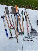 Photo shows 8 yard tools laid on concrete with grass in background. Includes sledgehammer, square shovel, hoe, axe, cultivator, leaf rake, garden pruner, and grass rake.