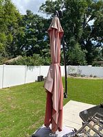 Side view of the large patio umbrella with closed red/pink canopy and black metal pole in a green backyard.