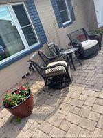Two dark wicker-style swivel chairs with cream cushions arranged outdoors on paved surface, small square table with glass top between them, large brown plastic flower pot with red flowers visible