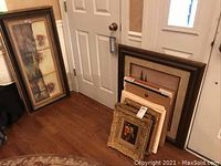Photo showing several framed paintings and artworks leaning against the walls, includes different sizes and frame styles.
