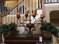 Full lot view showing rustic iron and tin candelabra on a wooden surface with four large pillar candles, flanked by two rectangular wicker baskets containing faux ivy and fern plants, positioned on either side of the candelabra.