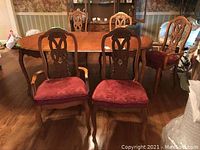 Five wooden dining chairs around a dining table, two captain chairs with armrests and three regular chairs without armrests. Upholstered red fabric seats. Carved wooden backrests with ornate patterns.
