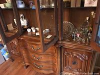 Photo of wooden display cabinet containing a variety of servingware items including silver plated bowl, brass utensils, decorative bottles, and set pieces visible through glass doors.