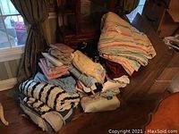 Large assortment of assorted pillows, folded blankets including striped and patterned comforters, and sheets stacked on a wooden floor near a window.