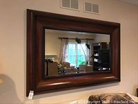 Full view of the large rectangular mirror mounted on a wall showing the dark wood thick frame with bevel glass reflecting a window and room.