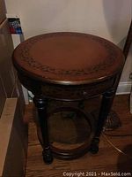 Full view of the round drum style accent table showing leather look crackled surface, wood legs and bottom shelf.