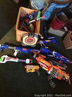 Photo showing the large basket filled with various Nerf guns and toys placed on a carpeted floor next to an upholstered chair and other household items.