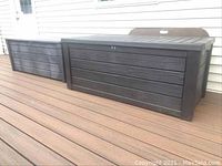 Two Keter outdoor deck storage boxes side by side on a brown wooden deck, showing weathered black resin material with horizontal panel texture, viewed from the front.