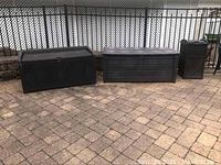Three outdoor units on a patio: two Keter deck storage boxes and one Sun Cast outdoor garbage bin, all black and weathered with surface sun fading.
