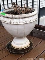 Front angled view of one cream-colored pedestal flower pot showing decorative leaf pattern near rim and base, weathered with dirt streaks and filled with soil and dry plant material