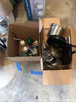 Two cardboard boxes; one contains various children's drink thermoses and NY Yankee cups, the other with pots and pans including lids and cookware items.