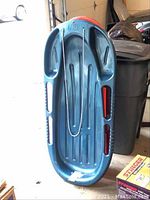 Blue plastic sled displayed upright showing bottom side with grooves and pull rope attached.