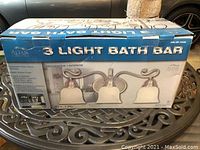 Photograph of the original blue and white box for the Altair 3-light bath bar, showing product image and branding.