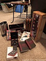 Overview of barware and wine accessory items set on carpet next to small table including flask, wine set, boxed books, and party basketball game.