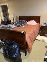 Full view of the queen size wooden sleigh bed frame with decorative orange bedding on it, situated in a carpeted room with nightstand and lamp beside it.