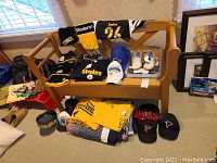 Photo showing a bench with multiple Pittsburgh Steelers apparel items, Steelers slippers, Falcons caps, folded yellow Steelers t-shirt, and football keychain.