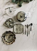 Vintage silver and glass serveware including relish bowl with spoon, butter dish, salt and pepper set, covered candy dish, table forks and spoons, all displayed on a white surface.