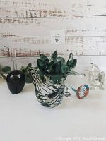 Photo showing vintage green decorative bowl, black glass cat figurine, clear glass squirrel, two birds (clear and multicolored), and clear glass slender vase or candle holder on a white surface, rustic wood background.