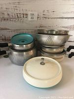 Cookware set showing 4 aluminum pots with black handles, metal colander, several baking pans stacked, and turquoise lid Dinex thermal bowl with white base.