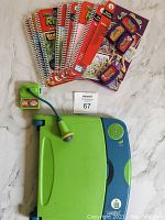 Green and blue LeapFrog LeapPad learning device with attached microphone and stylus, seven spiral-bound educational books and cartridges arranged on a white surface.