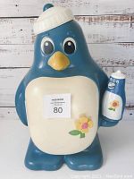Front view of large blue and white penguin fridge holding a small penguin teapot in its right hand, showing flower decoration on belly