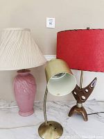 Set of three lamps: pink ceramic lamp with beige pleated shade, gold flexible neck desk lamp, retro ceramic lamp with wooden base and red shade.
