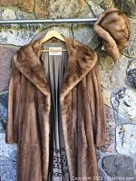 Front view of full-length brown mink fur coat with matching hat on hanger against stone wall.