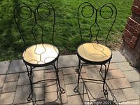 Front view of two wrought iron chairs with round yellow seats, showing decorative scroll backs.