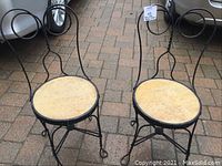 Front view of 2 wrought iron chairs with circular birch wood seats on a brick patio.