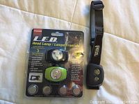 Three headlamps arranged on a white fabric background; two LED headlamps in packaging with button batteries, one green and black, and one Petzl black headlamp with elastic strap.