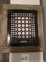 Top-down view of the assembled floor grate showing the decorative outer frame with heavy paint and the central black grate with geometric cutouts.